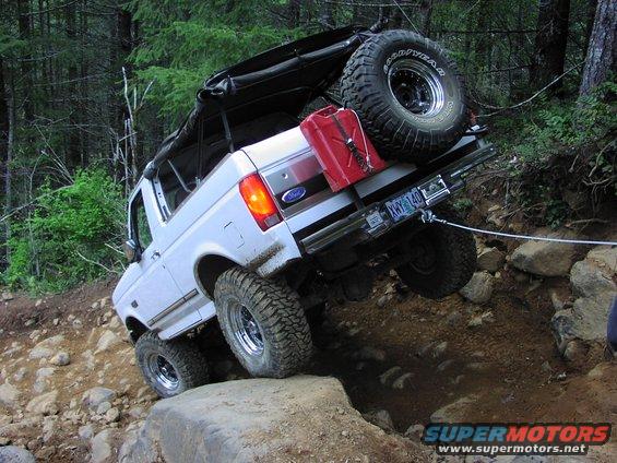 1993 Ford Bronco with soft top, spare tire, and gas can, climbing over rocks on a forest trail