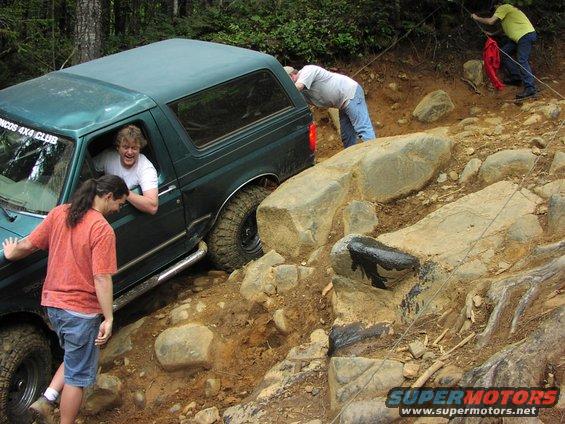 Green SUV navigating rocky terrain with people outside helping. Text on door reads 'BRONCOS 4X4 CLUB'