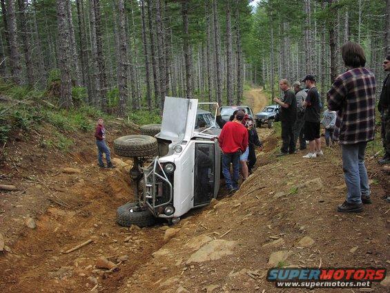White off-road SUV on its side in a ditch, hood up, with people gathered around