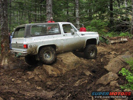 White SUV with a black top navigating a muddy, rocky trail with trees all around