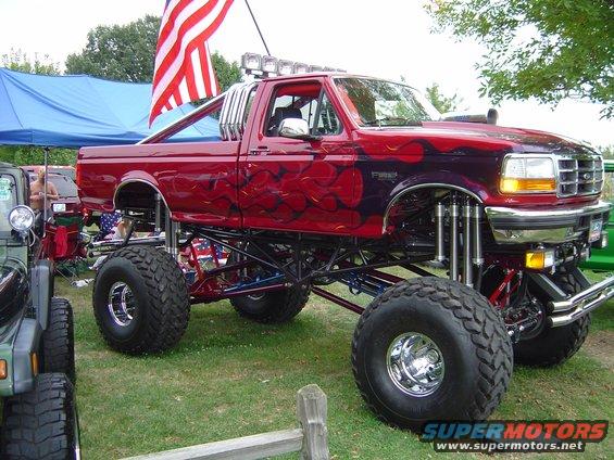 Lifted red pickup truck with flame decals and large tires, parked next to a green Jeep
