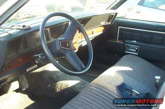 Car interior with blue steering wheel, wood-grain trim, and striped fabric seats. A "SUPERMOTORS" watermark is visible