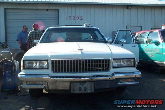 Front view of a white 1986 Chevrolet Caprice with its driver's door open. A teal taxi is parked to its right