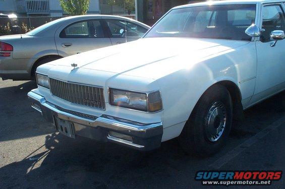 White Chevrolet Caprice front end, grille has 'CHEVROLET' text. Another car is parked behind it
