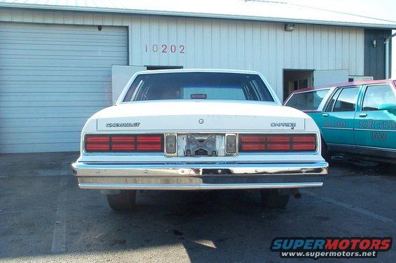 Rear view of a white 1980s Chevrolet Caprice sedan parked in front of a garage, with a teal taxi visible to the right