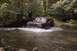 1994 Ford Bronco - A red SUV navigates a stream in a wooded area, kicking up water as it moves forward.