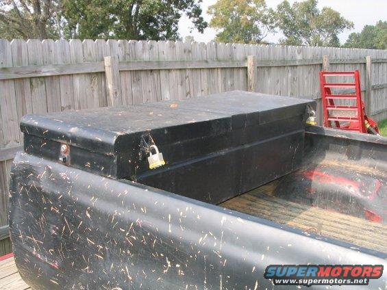 Black toolbox locked in a dirty truck bed with a wooden fence behind it