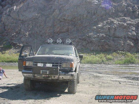 Dark-colored Ford Bronco II with off-road lights on roof, parked on dirt with a person sitting nearby