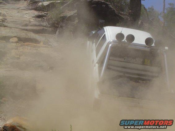 White SUV kicking up dust on a rocky incline, with a roll cage and auxiliary lights
