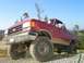 1989 Ford Bronco - This lifted red Ford Bronco is parked on a gravelly surface with trees and hills in the background.