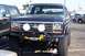 1990 Ford Bronco - A lifted dark blue Ford Bronco with a winch and auxiliary lights is parked in a lot.