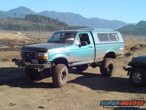 Modified blue pickup truck with a camper shell parked on a dirt road, with another vehicle partially visible to the right