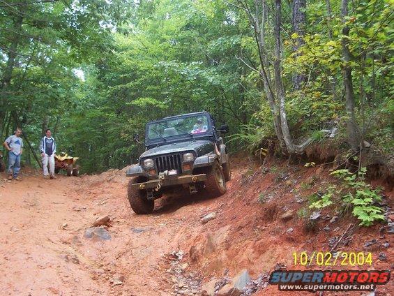 Jeep Wrangler TJ driving on a muddy trail with trees around. Two people and an ATV are visible in the background
