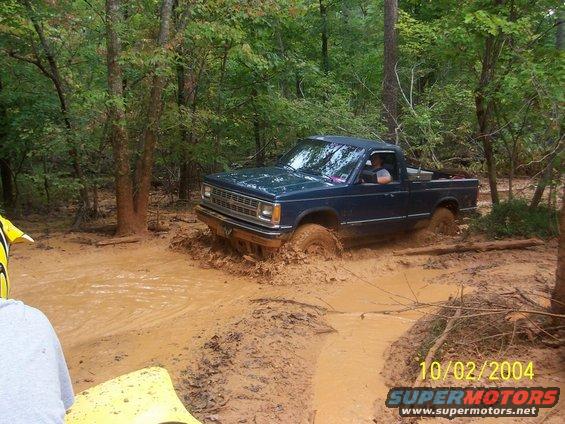 Blue pickup truck stuck in mud on a wooded trail. The front grille suggests an early 1980s model