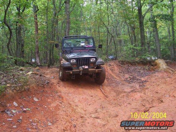 Jeep Wrangler driving on a muddy trail in woods. Another Jeep is visible in background
