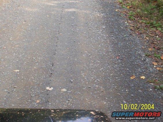 Gravel road with a dark vehicle fender in the foreground. Date stamp reads 10/02/2004