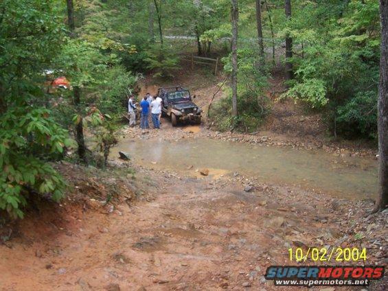 1994 Jeep YJ navigating a muddy off-road trail with people standing nearby. Another vehicle is partially visible in the background