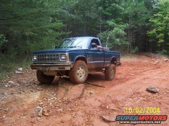 Blue pickup truck driving on a muddy, rocky trail in the woods