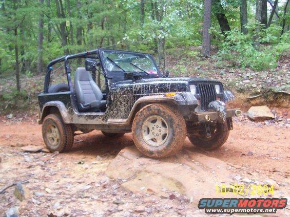 Black Jeep Wrangler on a dirt trail, covered in mud. Front 3/4 view