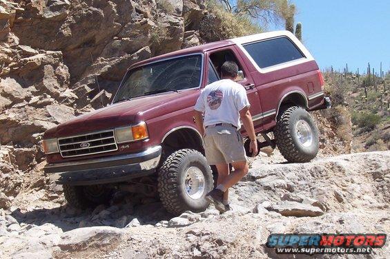 Maroon 1990 Ford Bronco navigating rocky terrain with a person walking beside it