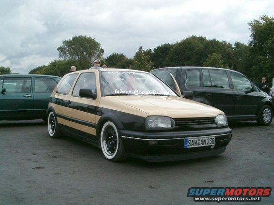 Tan and black VW Golf Mk3 with whitewall tires and a sticker on the windshield
