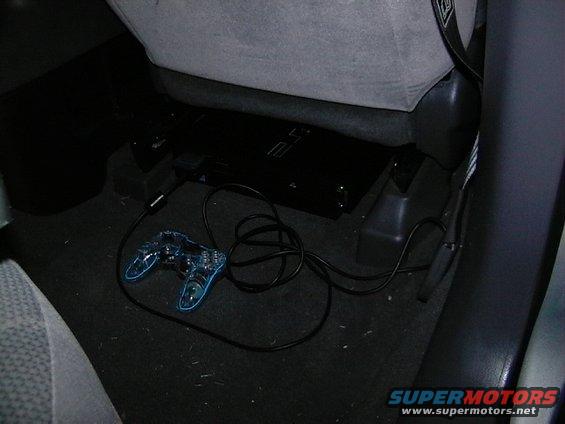 Black PlayStation 2 console and transparent blue controller on car floor, connected by wires
