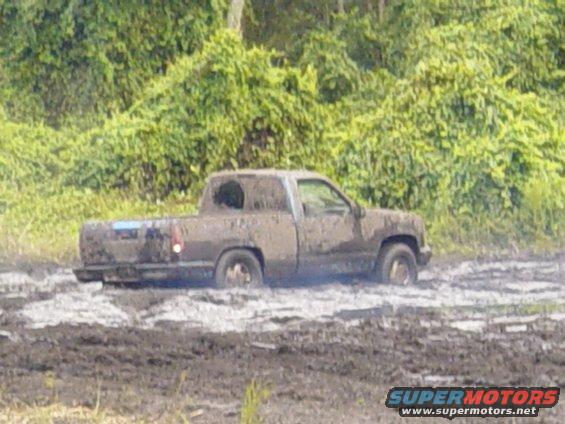 Mud-covered pickup truck stuck in mud, side view. Visible taillight and part of front fender