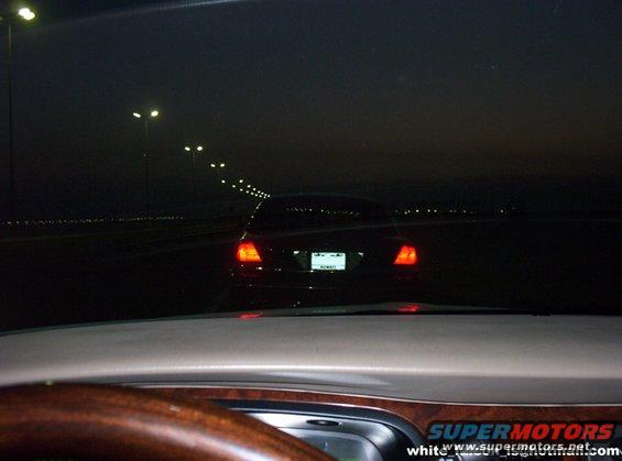 Car driving on a dark road at night, with streetlights in the distance. The taillights of the car ahead are visible