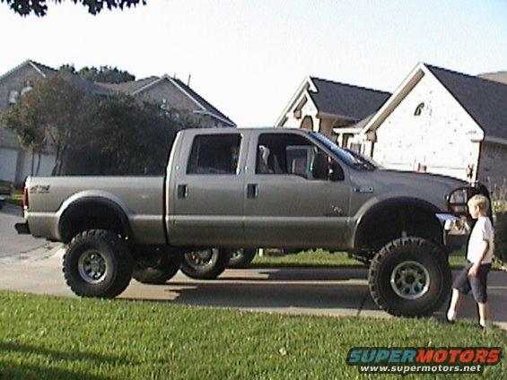 Tan lifted Ford F-250 crew cab on big mud tires in a driveway with a child standing nearby