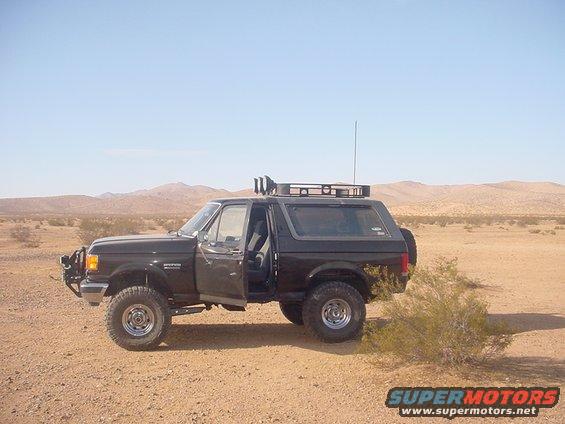 Black Ford Bronco with open driver door, roof rack, and aftermarket bumper parked in a desert landscape