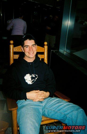 Man in a black hoodie and blue jeans sits in a wooden rocking chair, hands clasped