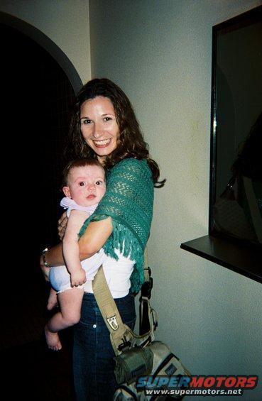 Woman holding a baby. She has long brown hair and is wearing a green shawl over a white shirt and jeans. A bag is slung over her shoulder