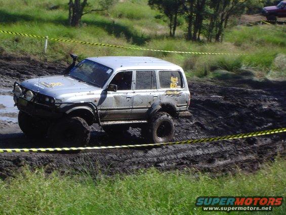 Silver Toyota Land Cruiser going through mud with large tires and a snorkel. Yellow caution tape is visible