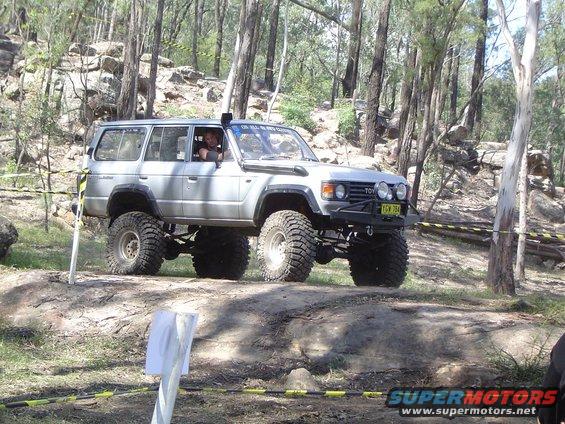 Silver off-road SUV with large tires and a roll cage, navigating a wooded trail