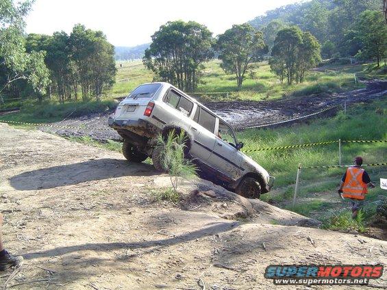 White SUV tilted on a rocky off-road trail with mud pits in the background. A person in an orange vest watches