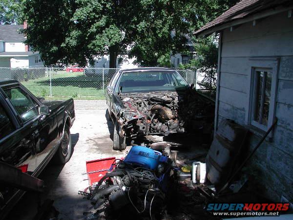 Car engine bay with disassembled parts and a black car to the left