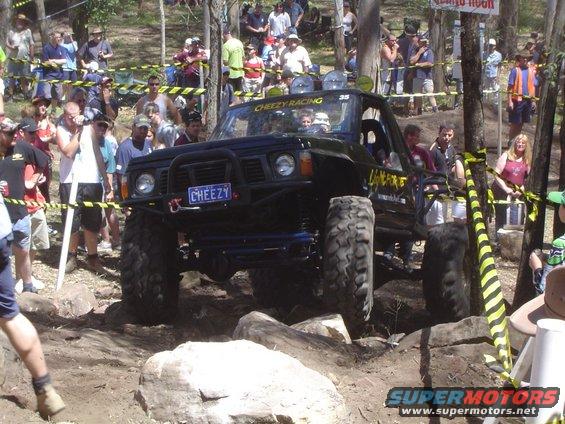 Modified off-road vehicle with large tires navigating rocky terrain, crowd watching