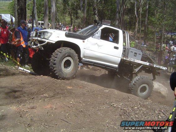 White off-road pickup truck with large tires climbing a dirt hill, snorkel, and custom flatbed