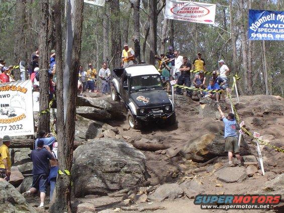 Black off-road vehicle with a white top, climbing rocks, with spectators watching