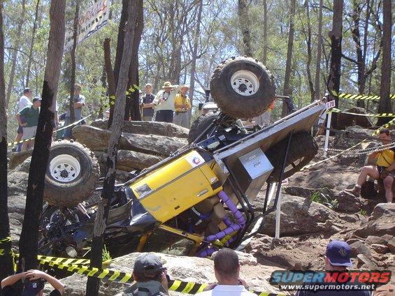 Yellow off-road vehicle overturned on rocks during an event, with spectators watching