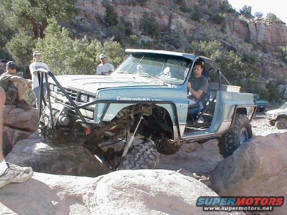 Light blue modified pickup truck with off-road tires navigating rocks. "ColoradoKS.com" text visible on door