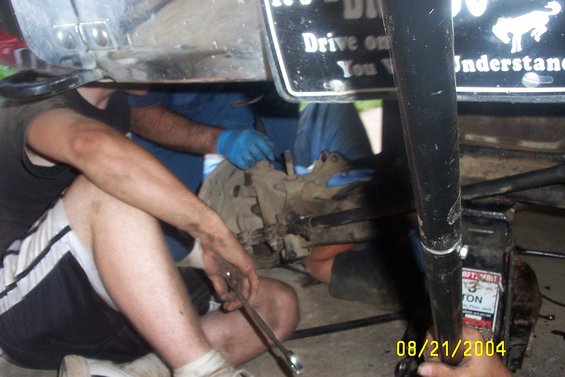 People working on the underside of a vehicle, possibly a truck, with tools and a jack visible. A license plate with text is mounted above
