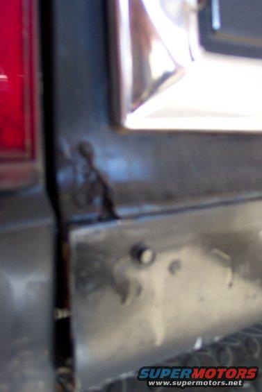 Close-up of rust on a vehicle's tailgate corner, showing a red taillight on the left