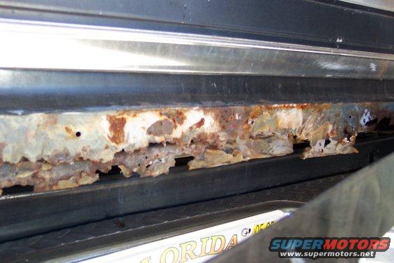 Heavily rusted rocker panel on a vehicle, showing significant metal decay and holes. Part of a Florida license plate is visible below