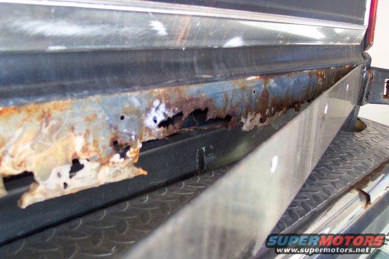 Close-up of heavily rusted lower body panel on a vehicle. Shows significant corrosion and holes