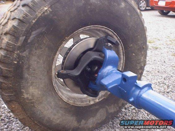 Close-up of a blue front axle assembly, tire, and brake rotor. Two other vehicles are visible in the background