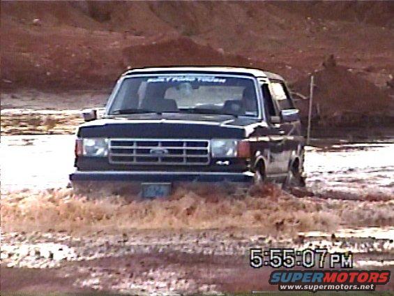 1988 Ford Bronco driving through muddy water, front view. 'Built Ford Tough' sticker on windshield