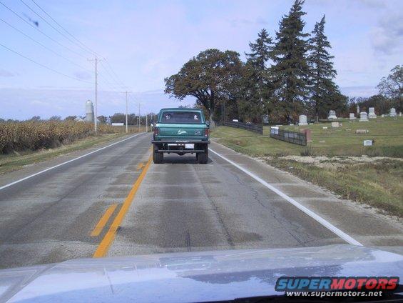 Green pickup truck seen from behind on a two-lane road, with a cemetery on the right