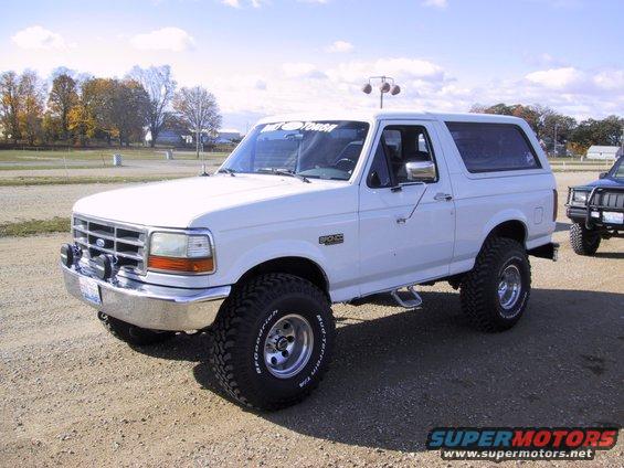 White Ford Bronco parked on dirt lot, another SUV in background. Has a 'MOTOR' sticker on windshield