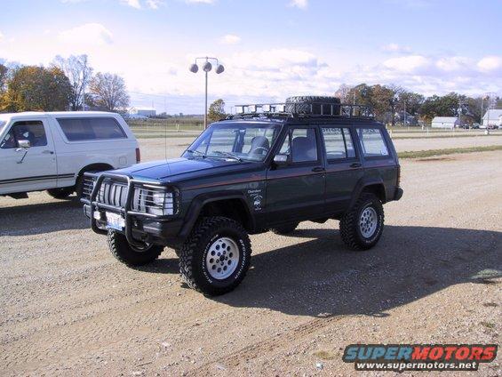 Dark green Jeep Cherokee with a roof rack and spare tire, front bull bar, and aftermarket wheels